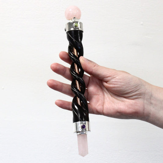 A hand holds a crystal wand, featuring a twisted black shaft with rose quartz end pieces and a silver-colored metal setting, against a plain white background.