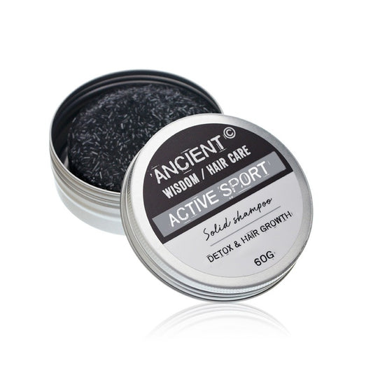 Round metal tin of solid shampoo sits open revealing a black shampoo bar inside on a white reflective surface, label reads "ANCIENT © WISDOM / HAIR CARE ACTIVE SPORT Solid shampoo DETOX & HAIR GROWTH 60G."