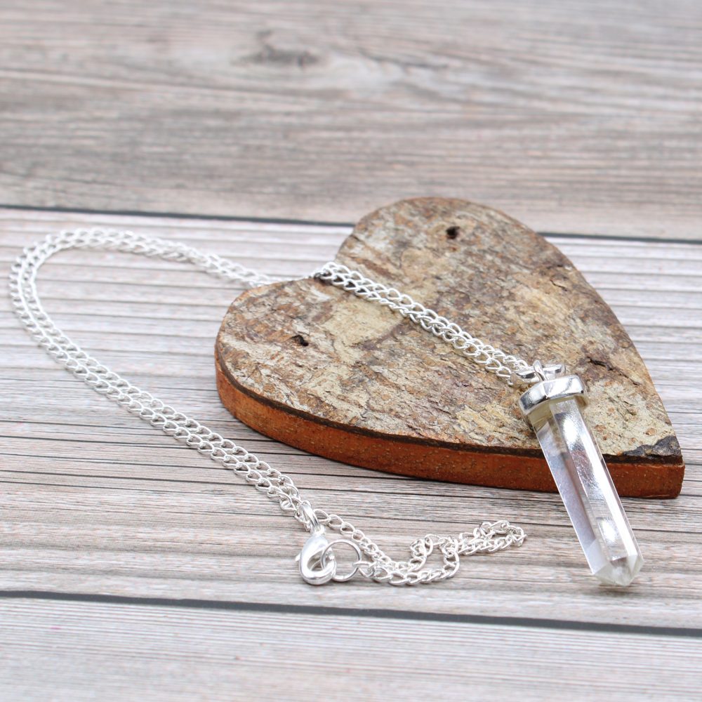 A clear, crystal-like pendant with a silver chain lies atop a small, heart-shaped wooden block. The block rests on a light-colored wooden surface.