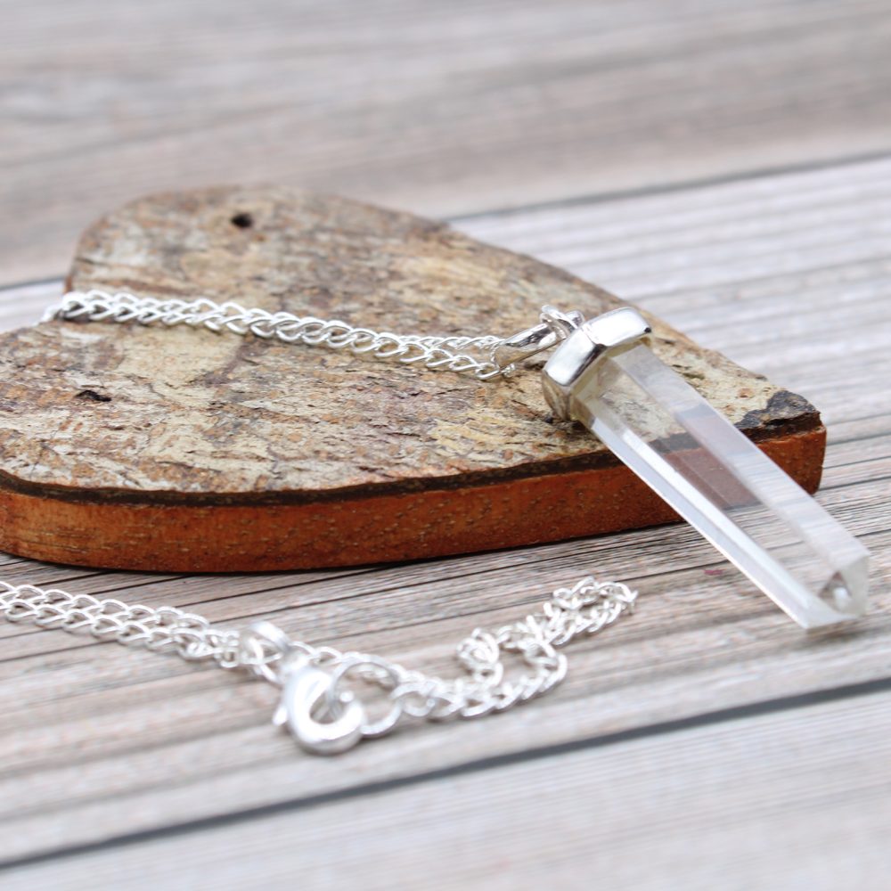 A crystal point pendant with a silver chain lays across a wooden heart on a light wooden surface.