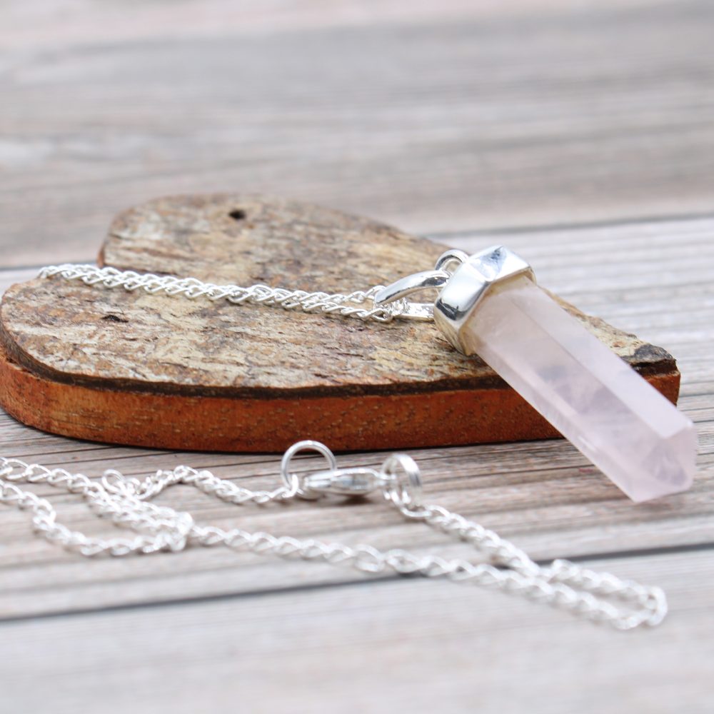 A quartz crystal necklace rests on a small, rough-hewn wooden platform. The necklace chain drapes over the platform and onto a weathered, wooden surface.
