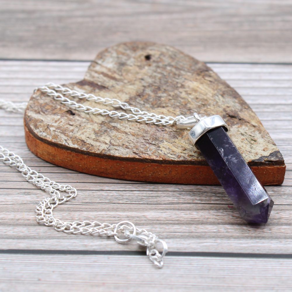 A silver chain necklace with an amethyst point pendant rests diagonally across a weathered wooden heart on a wooden surface.