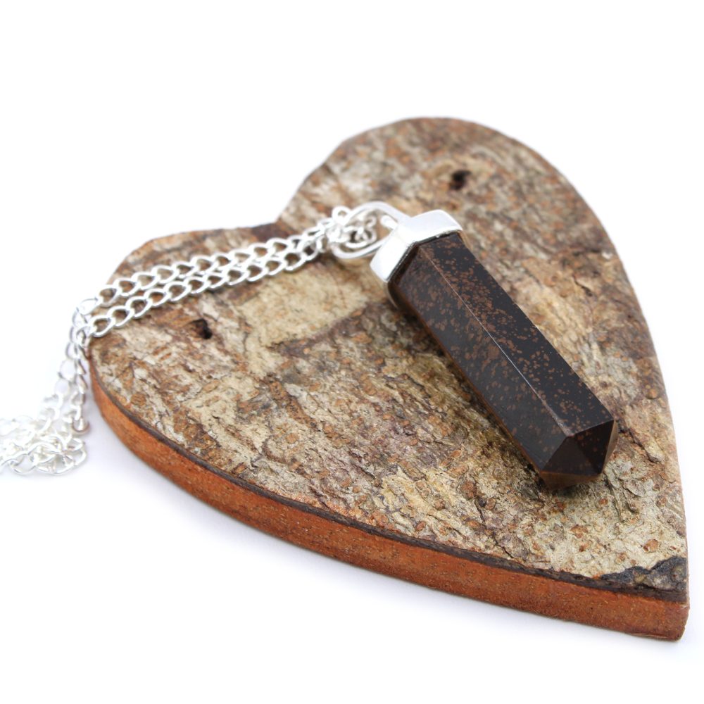 A dark, hexagonal crystal pendant rests on a silver chain atop a heart-shaped piece of tree bark, which is displayed against a white background.