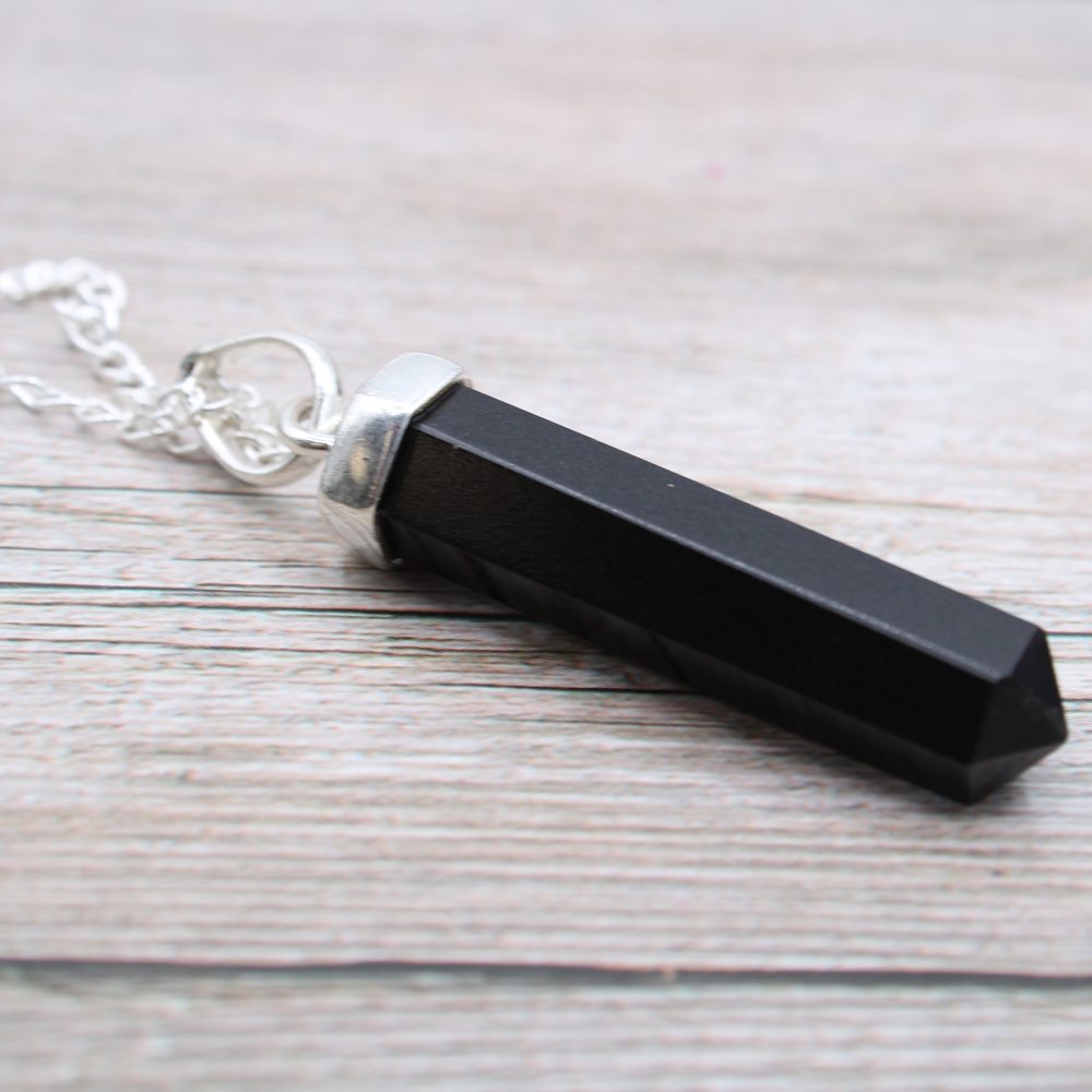 A black, hexagonal crystal point pendant hangs from a silver chain. It lies on a background of pale, weathered wooden planks.