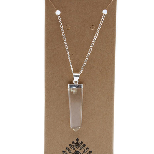 A clear, pointed crystal pendant hangs from a silver chain, displayed on a brown cardboard tag. An artistic logo, resembling leaves and a pyramid, is visible at the tag's bottom.