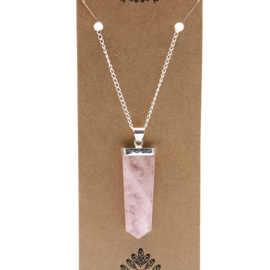 A rose quartz crystal pendant hangs on a silver chain. The pendant is displayed on a light brown cardboard backing with a leafy pattern at the bottom.