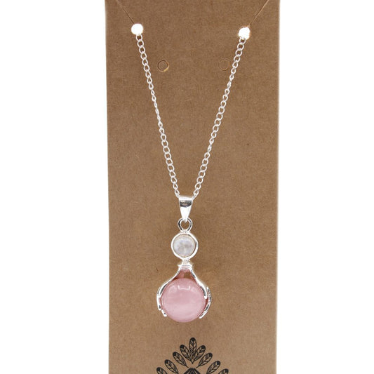 A silver chain necklace, with a light pink, spherical pendant stone nestled in a silver cage, hangs against a brown cardboard backing. Above the pink pendant stone is a smaller, white stone.