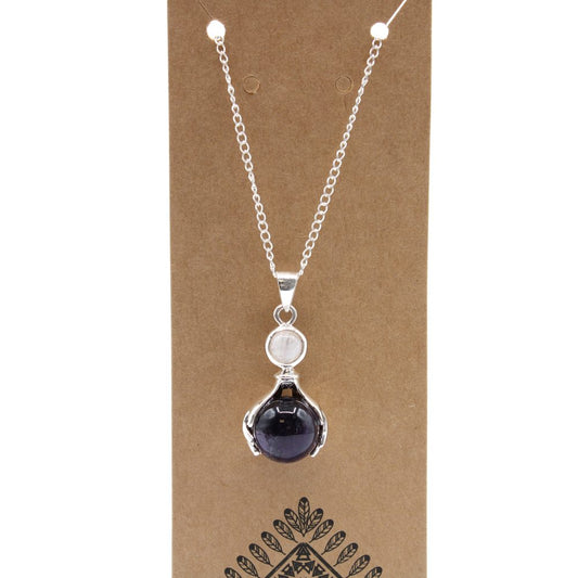 A silver chain necklace hangs on a cardboard display. The pendant has a dark purple stone held by silver claws under a smaller, white stone. The display features a symmetrical plant pattern at the bottom.