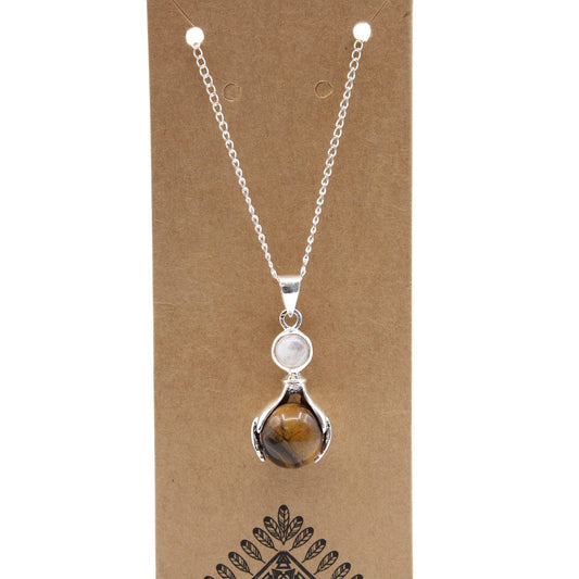 A necklace is displayed on a brown cardboard background. It has a silver chain and a pendant with a brown sphere nestled in silver arms under a milky white bead. Below the pendant are symmetrical leaf-like designs.