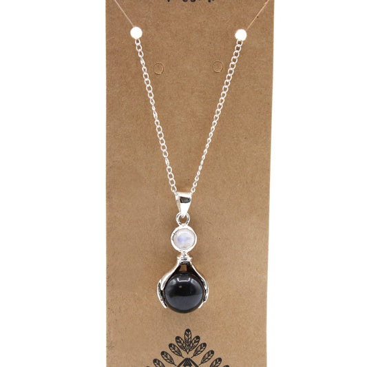 A silver chain necklace displays a black sphere and white moonstone pendant against a brown, rectangular display card decorated with a stylized leaf pattern.