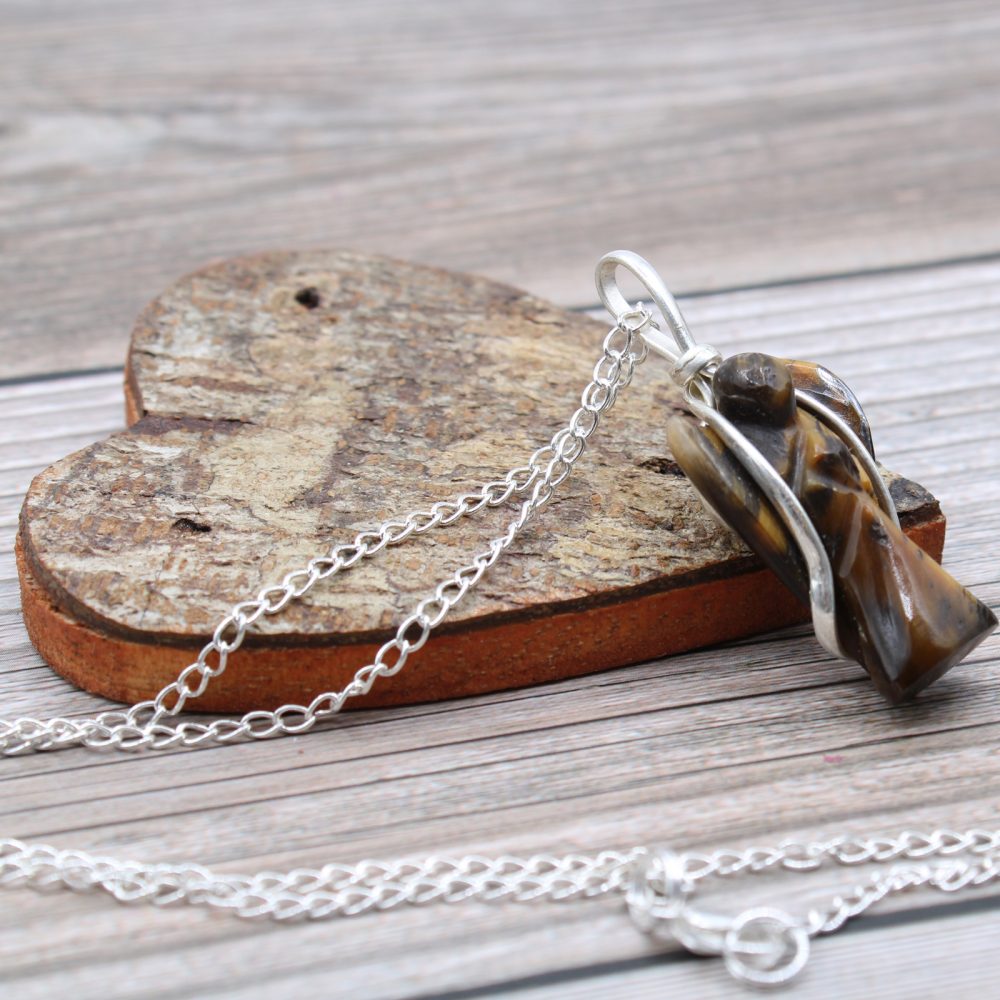 A tiger's eye angel pendant with a silver chain rests on a wooden heart against a wooden surface.