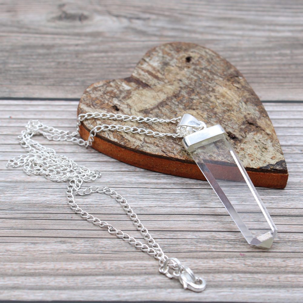 A quartz crystal point pendant on a silver chain necklace sits beside a wooden heart, all against a wooden background.