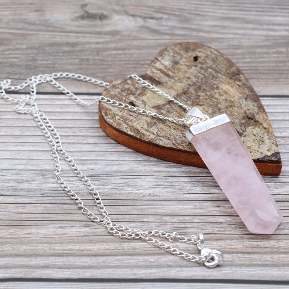 A pink, hexagonal prism pendant with silver hardware and a silver chain rests on a rustic, heart-shaped wooden surface with visible wood grain, which is set on a background of wood planks.