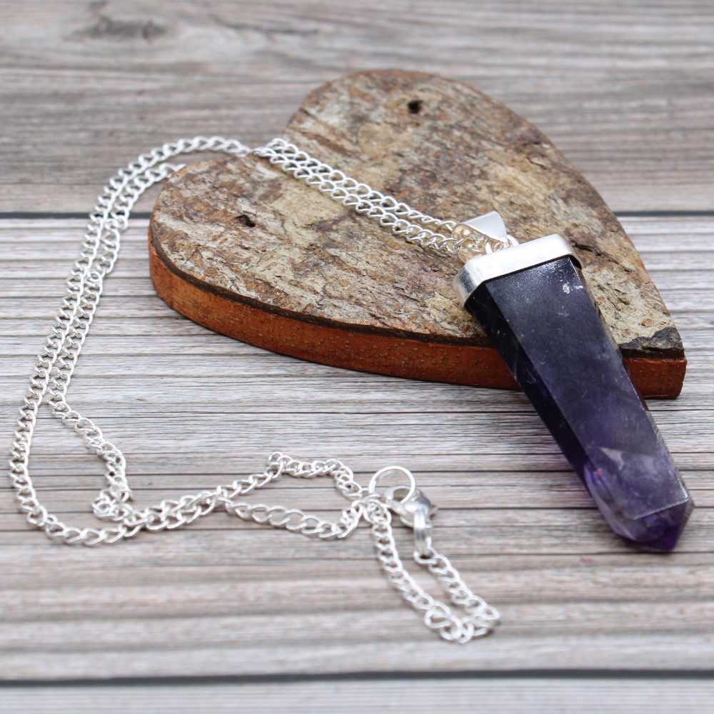 A purple crystal point pendant with silver chain rests on a heart-shaped piece of wood on a rustic, horizontally-oriented wooden surface.