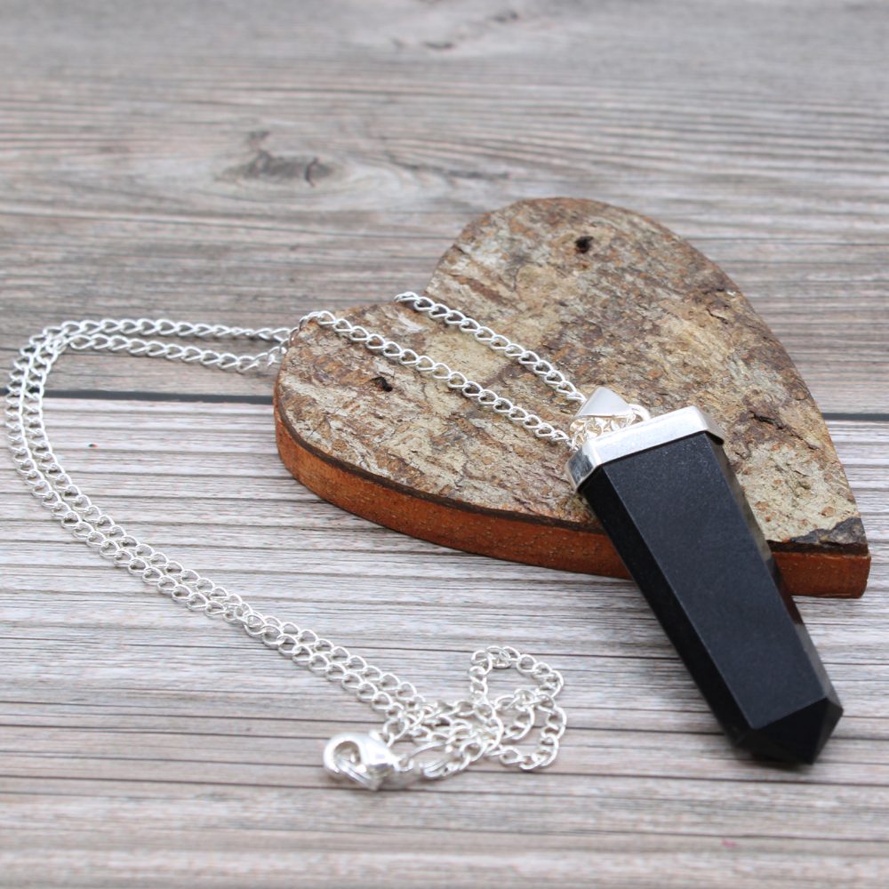 A black, silver-capped obsidian crystal pendant lies on a silver chain resting on a heart-shaped, wooden coaster against a light-colored wooden surface.