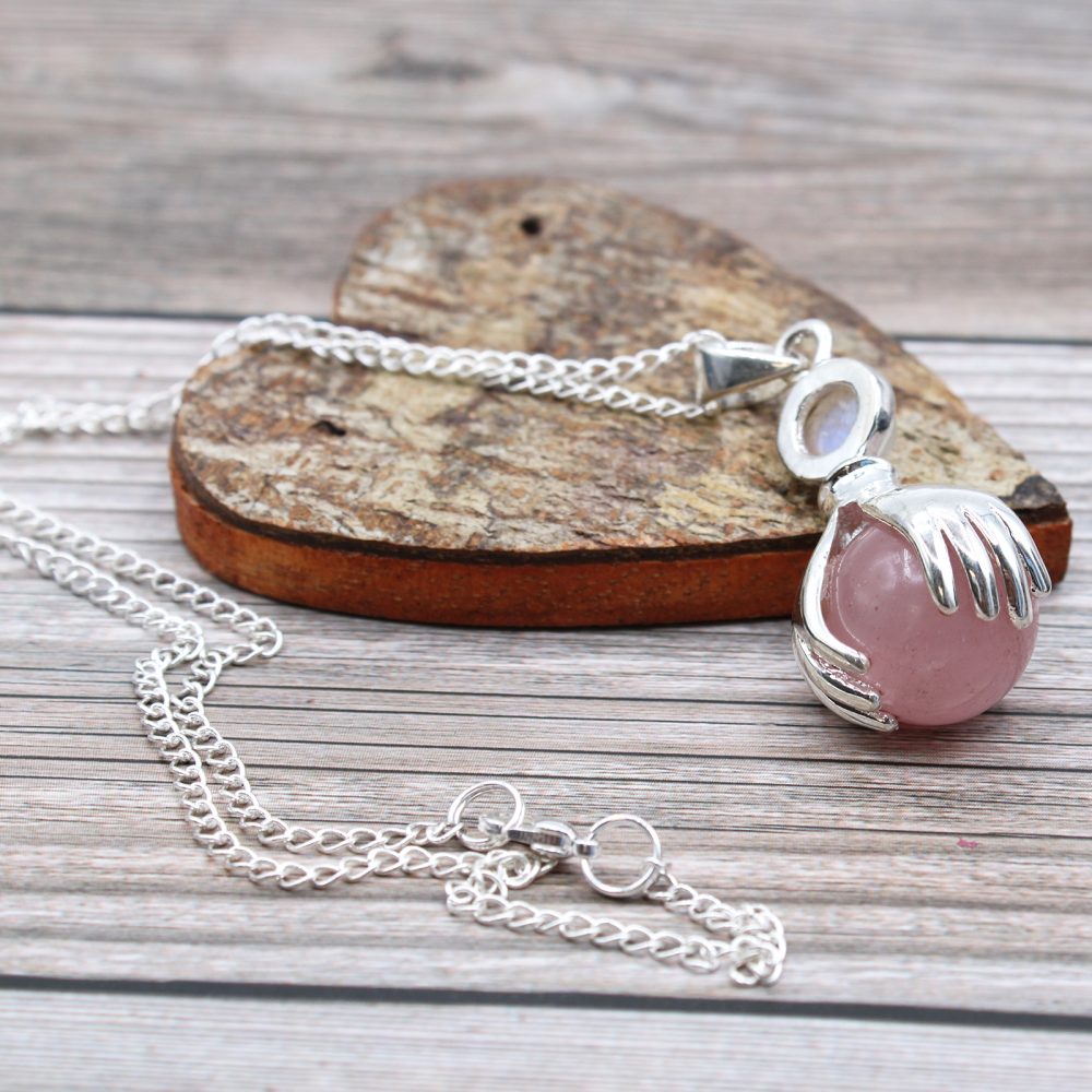 A silver chain holds a rose quartz orb necklace in a hand-shaped cage, presented on a wood slice. The arrangement rests on a light wood-grained surface.