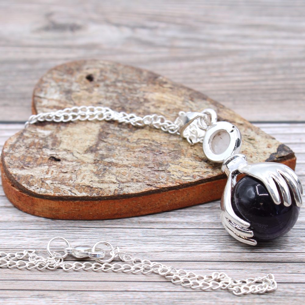 A silver hand-shaped pendant clasps a dark purple sphere, attached to a chain. The chain lies over a heart-shaped, rough-hewn piece of wood, all on a wood-patterned surface.