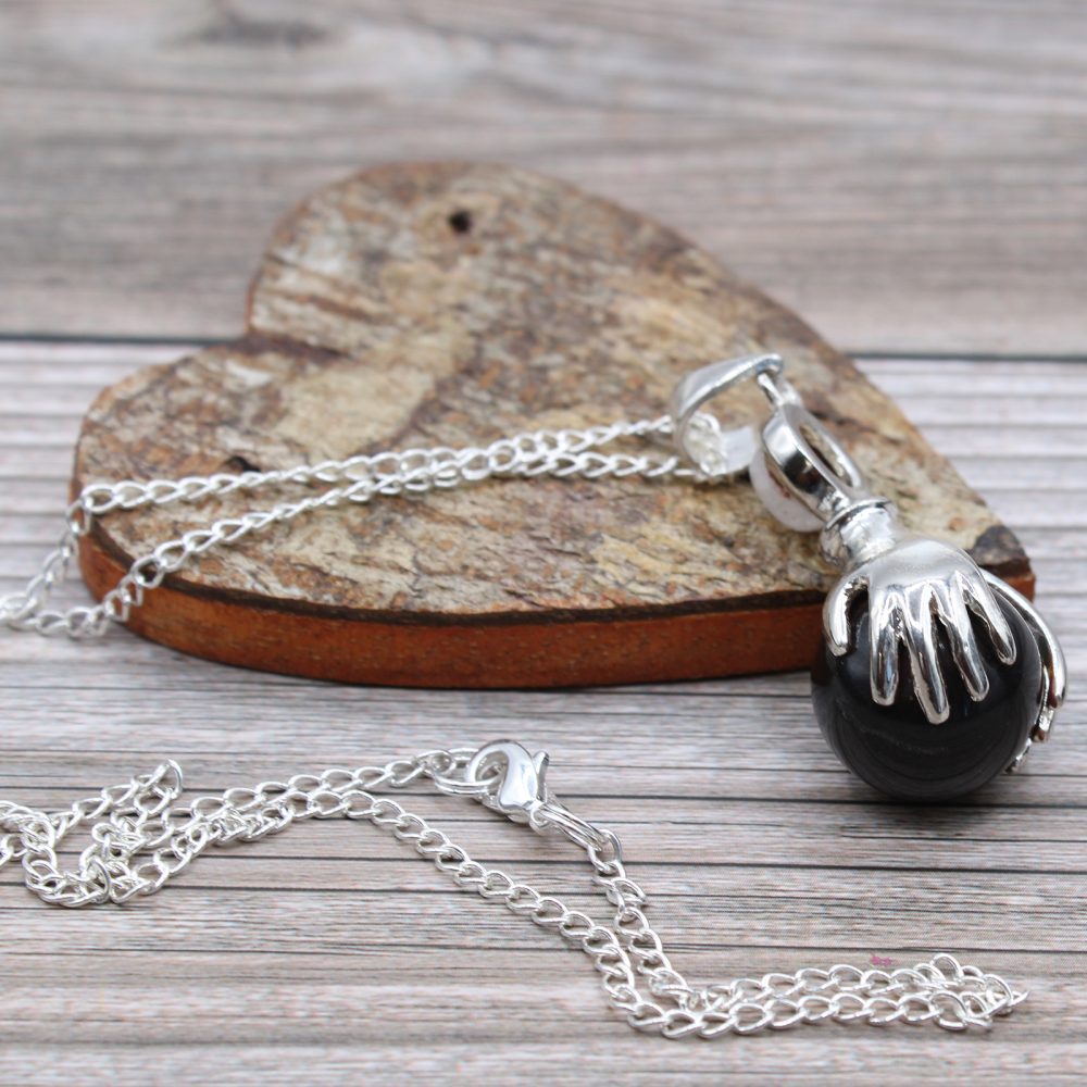 A silver necklace with a pendant resembling a hand holding a black orb sits on a wooden heart, which is in turn on a wooden surface.