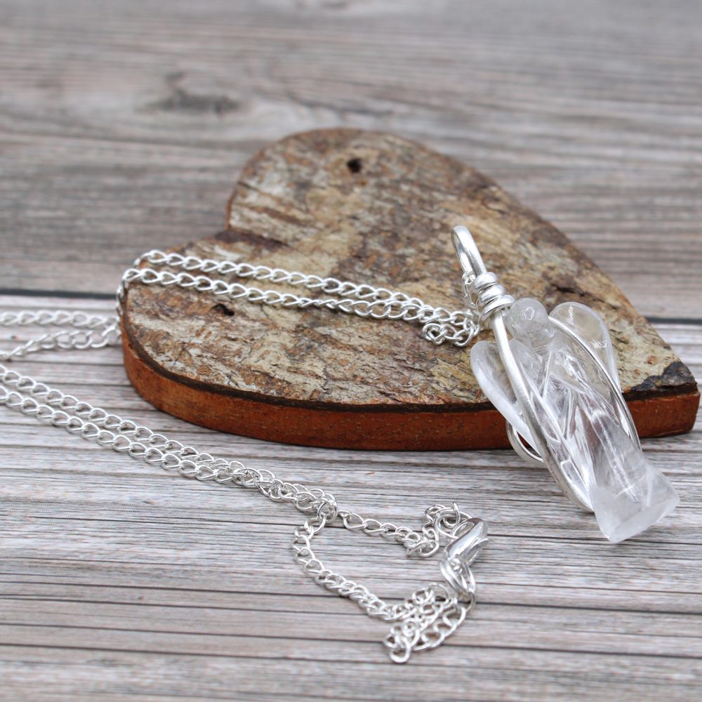 A crystal angel pendant, wrapped in wire and connected to a silver chain, lies on a rustic wooden heart atop a wooden surface.