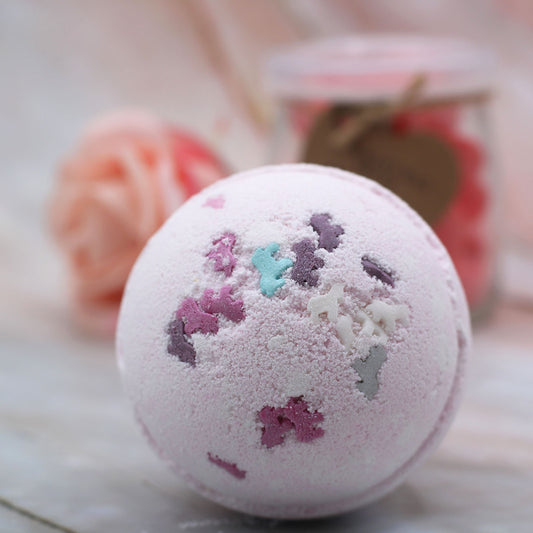A pink bath bomb adorned with multicolored star-shaped decorations sits on a marble surface. In the background, a blurred glass jar with a heart-shaped tag and a rose-shaped item create a delicate scene.