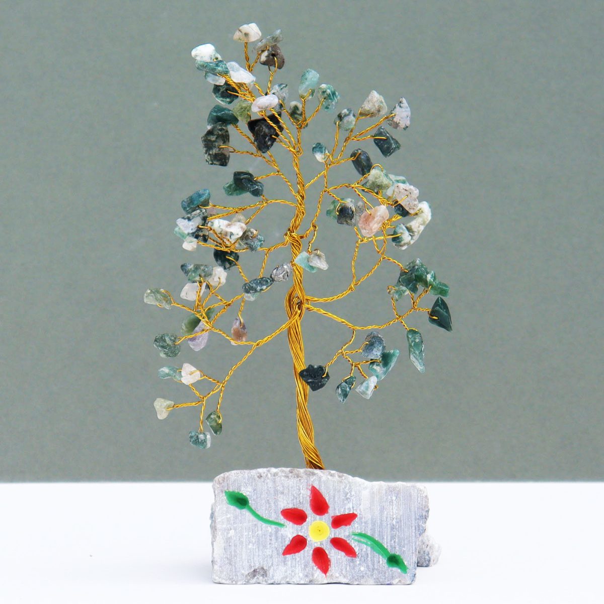 A small, decorative tree with golden wire branches bearing small, colorful stones, sits on a rectangular rock base painted with a red and yellow flower with green leaves. The background is a pale grey.