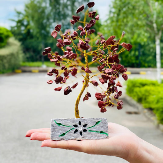 A hand presents a wire tree sculpture with gemstone leaves, set in a decorated stone base with a black flower design. The backdrop features a bright, blurry outdoor scene with greenery.