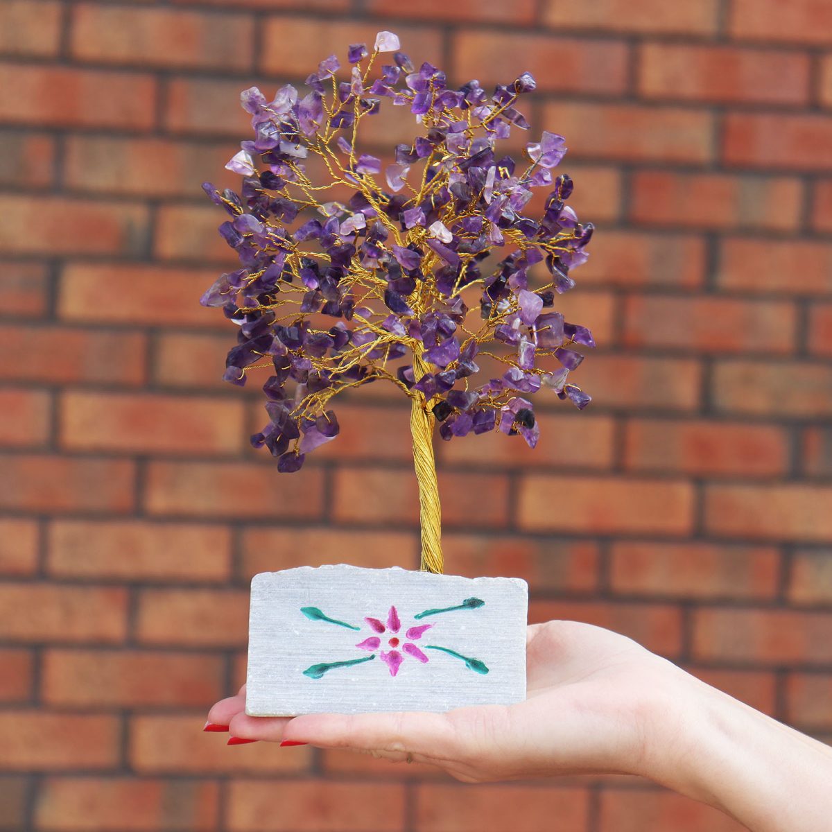 A small crystal tree with purple stones for leaves stands in a gray stone holder, which has a pink flower painted on it. The tree sits on a palm, against a brick wall.