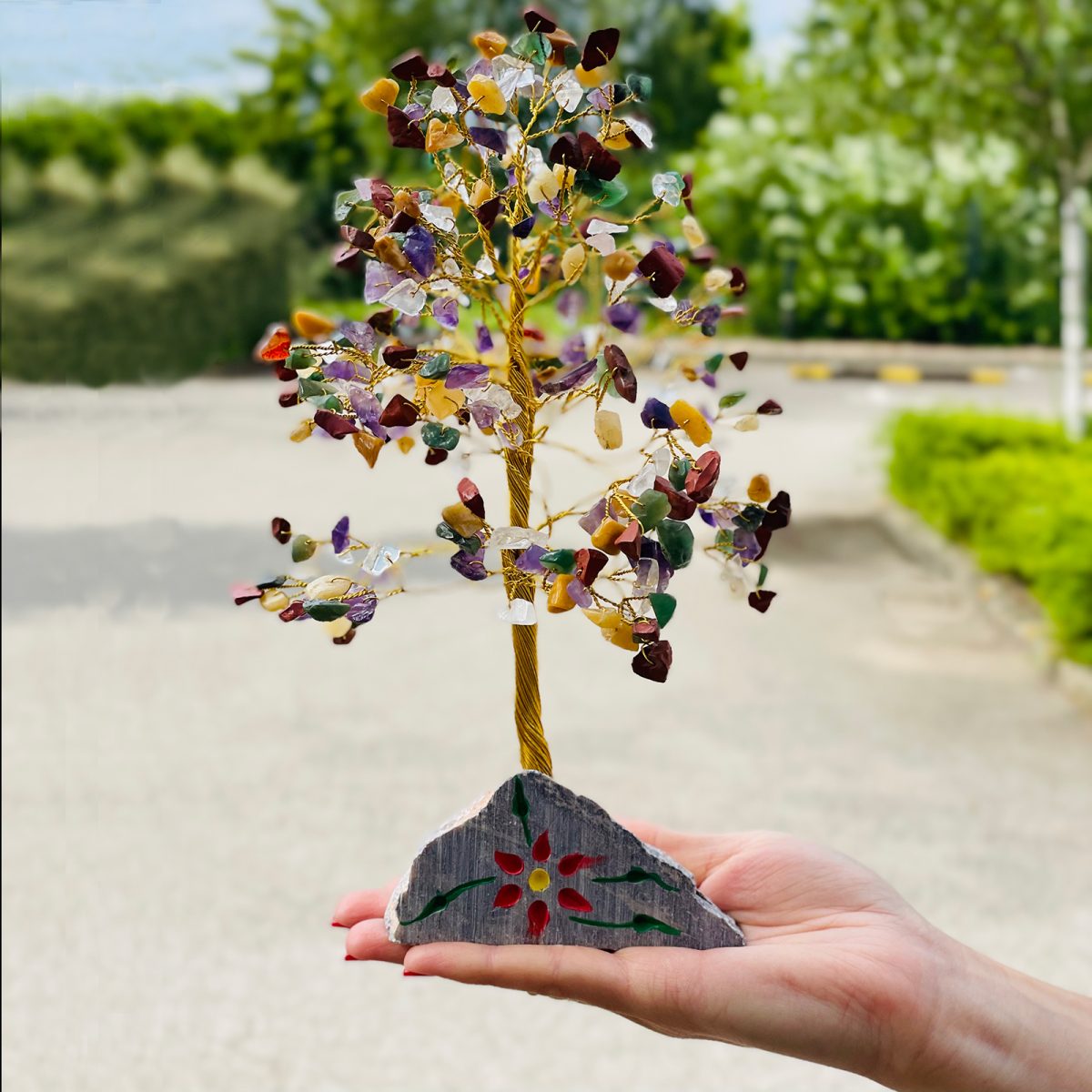 A gemstone tree is being held in a palm. The tree's leaves are small, colorful gemstones, its trunk gold wire, and its base a gray rock with a red and yellow flower. The background is blurred trees.