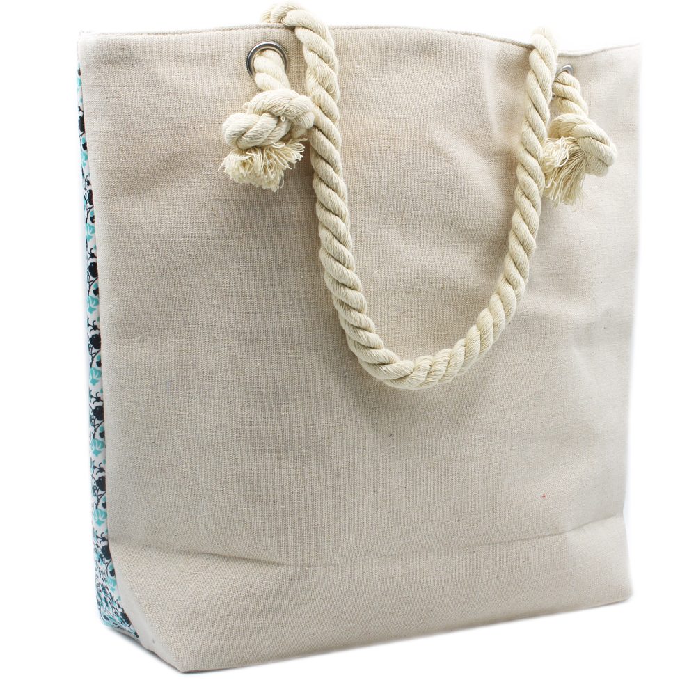 A beige canvas tote bag stands upright on a white surface. Rope handles attach to the bag through metal grommets, and a patterned blue fabric is visible along one side.