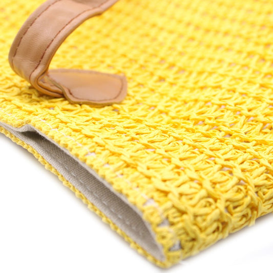 A woven, bright yellow handbag with a tan leather handle sits on a white surface. The woven texture is visible, and the lining is a light beige.
