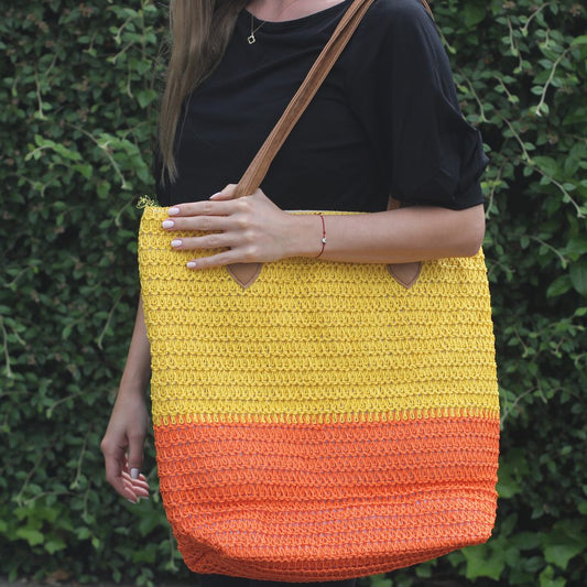 A woman carries a large, two-toned straw tote bag over her shoulder. The bag is half yellow, half orange. She stands against a green leafy hedge.