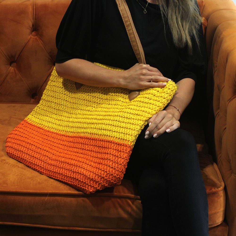 A woman sits, holding a straw tote bag with orange and yellow colors, on an orange tufted couch.