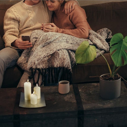 Couple cuddles on a sofa under a blanket, one holding a phone and mug; three lit candles and a potted plant rest on a low table in a living room.