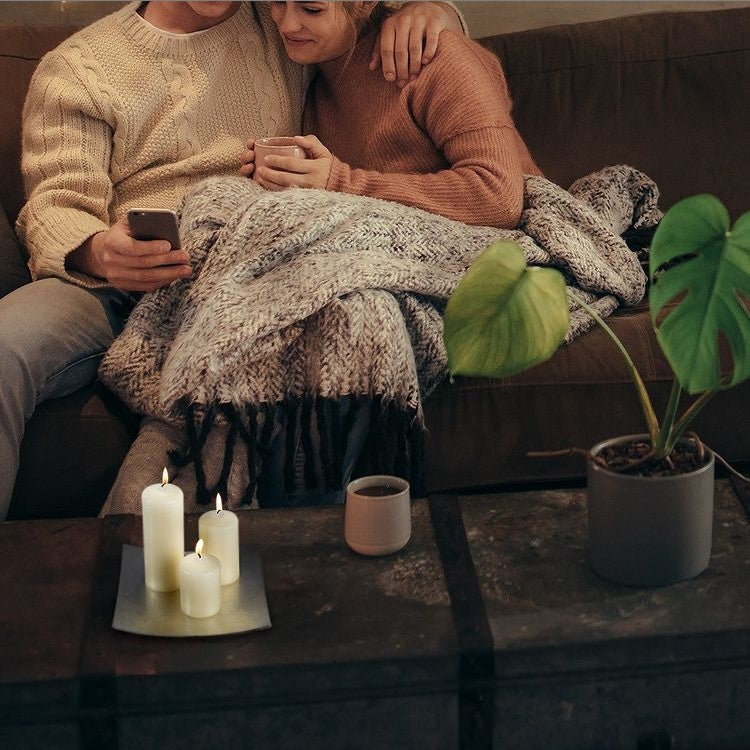 A couple sits under a blanket on a sofa. The man holds a smartphone, while the woman holds a mug. Candles, a plant, and a coffee mug sit on a coffee table.
