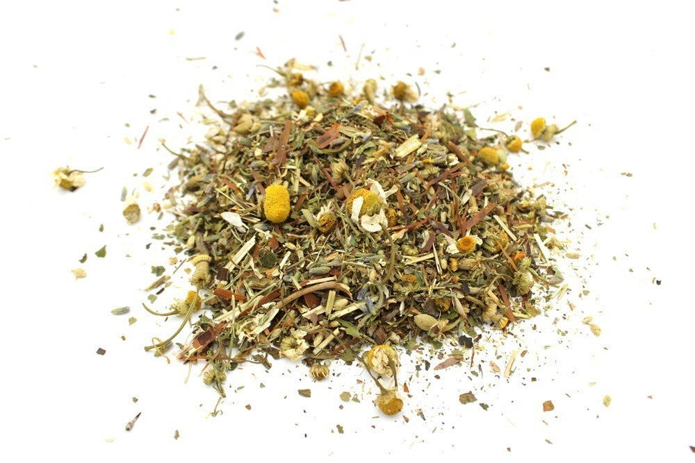 A pile of dried herbal tea blend with chamomile flowers is scattered across a white surface. This mixture includes various leaves, seeds, and stems, creating a colorful, textured appearance.
