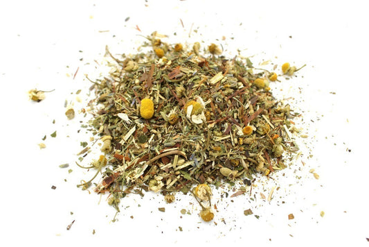 A pile of dried herbal tea blend with chamomile flowers is scattered across a white surface. This mixture includes various leaves, seeds, and stems, creating a colorful, textured appearance.