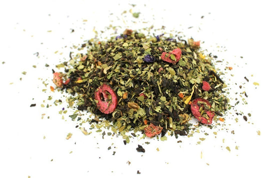 A pile of mixed loose tea leaves, including green herbs and dried red berries, lies scattered on a plain white surface, suggesting an aromatic and flavorful blend.