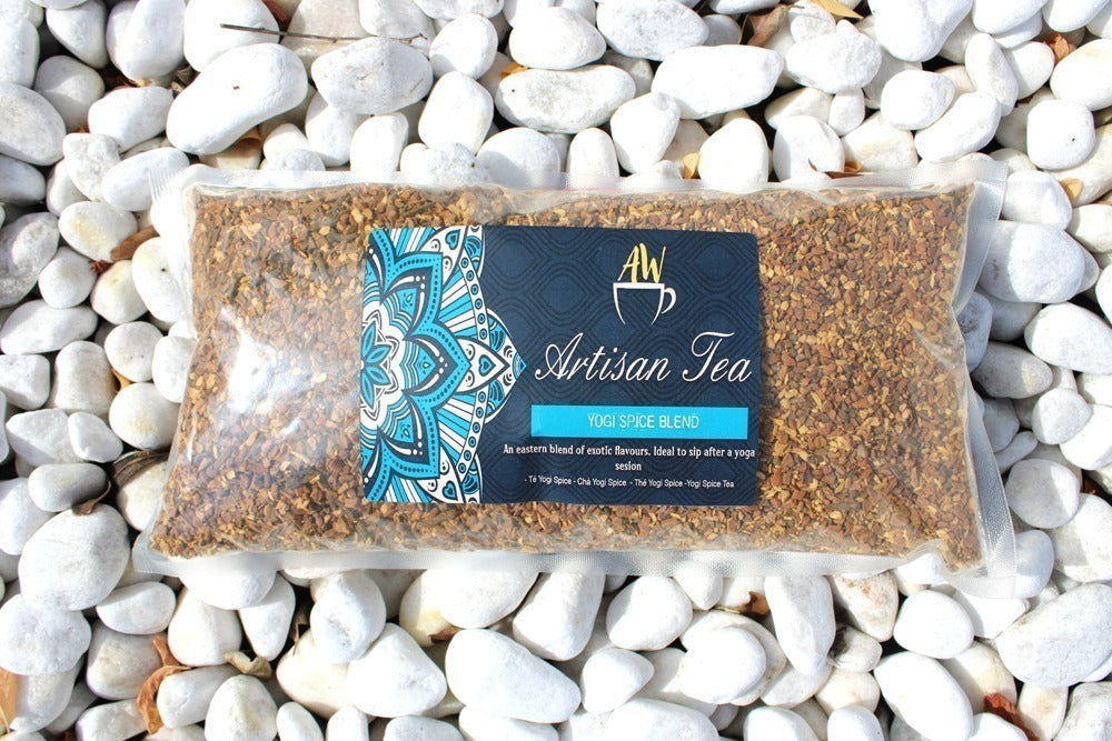 A plastic pouch filled with brown tea leaves labeled "AW Artisan Tea," "Yogi Spice Blend," rests on white pebbles. The package features a blue mandala design. Text: "An eastern blend of exotic flavours. Ideal to sip after a yoga session - Té Yogi Spice - Chaï Yogi Spice - Thé Yogi Spice - Yogi Spice Tea."