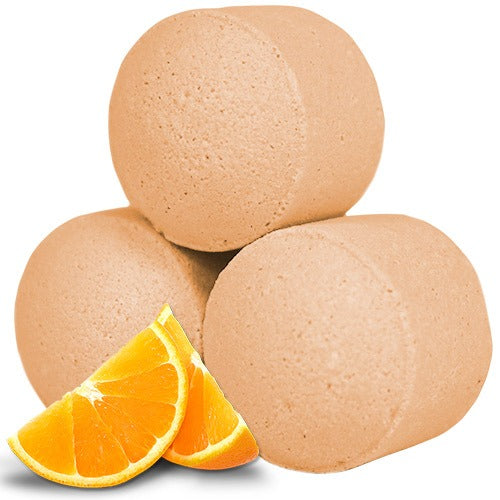 Three round, beige bath bombs are stacked in a pyramid shape, accompanied by slices of a vibrant orange fruit in the foreground. The background is plain.