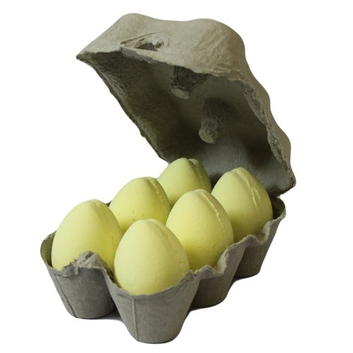 A gray egg carton holds six pastel yellow egg-shaped bath bombs, arranged neatly with the lid open, against a plain white background.