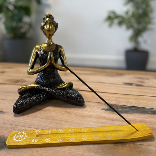 A golden figure sits cross-legged in a meditation pose with hands together. In front, a burning incense stick rests on a yellow holder. Background includes potted plants and a wooden surface.