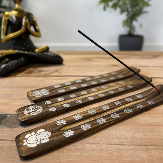 Wooden incense holders with decorative white symbols support a lit incense stick, releasing smoke. In the background, a meditative statue sits on a wooden surface, with a potted plant nearby.