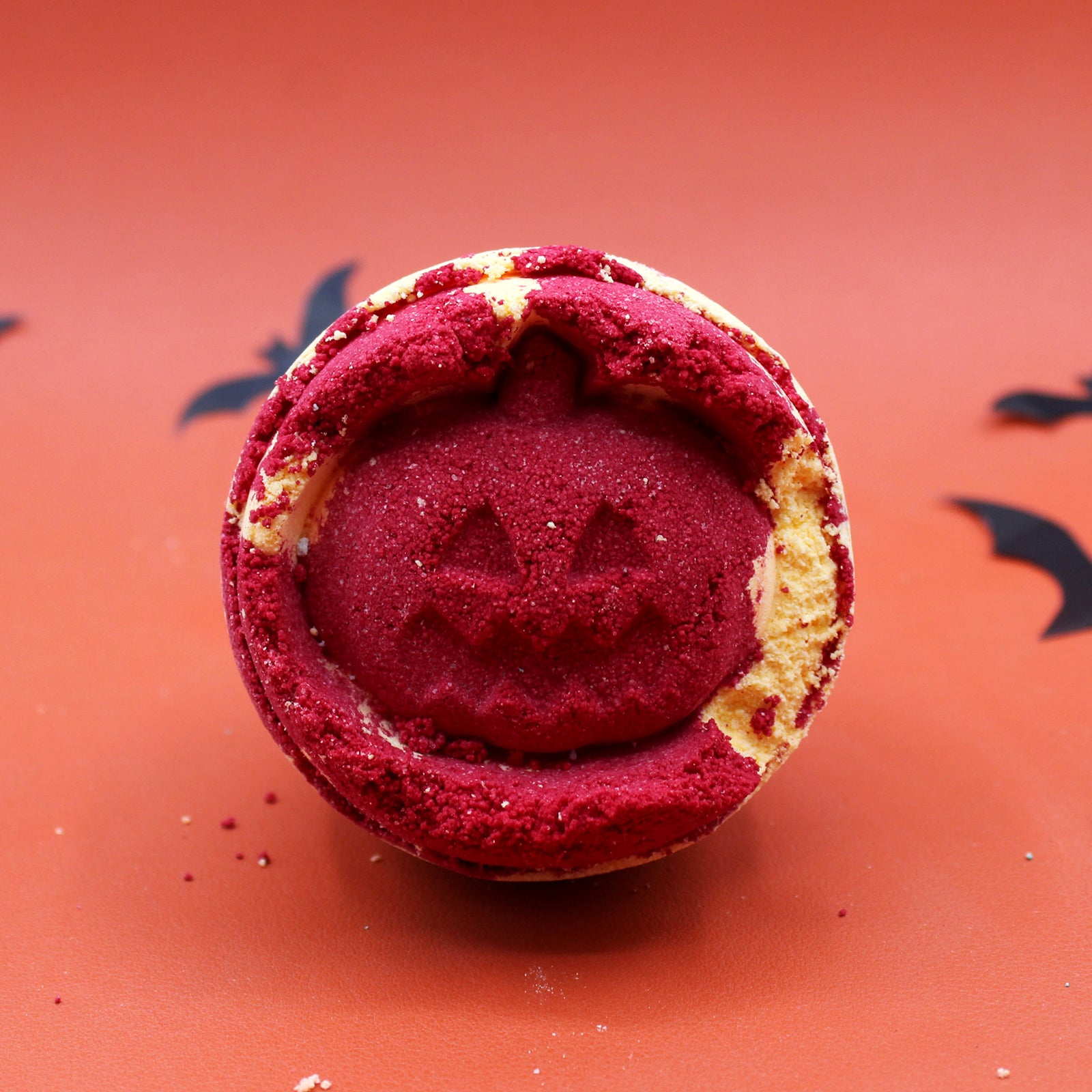 A red macaron features a carved jack-o'-lantern design. It rests on an orange surface with scattered black paper bats, creating a Halloween-themed scene.