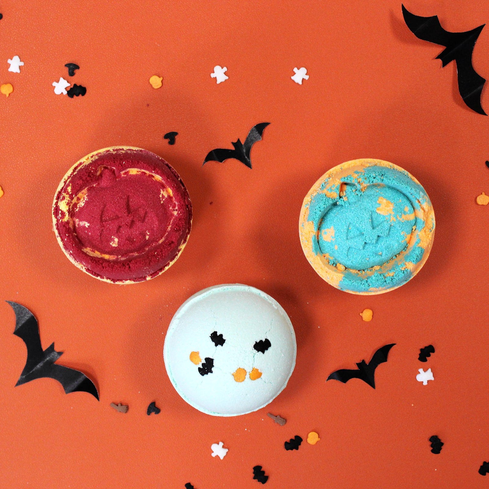 Three decorated macarons rest on an orange surface, surrounded by black bat and small Halloween-themed confetti, creating a festive Halloween atmosphere.
