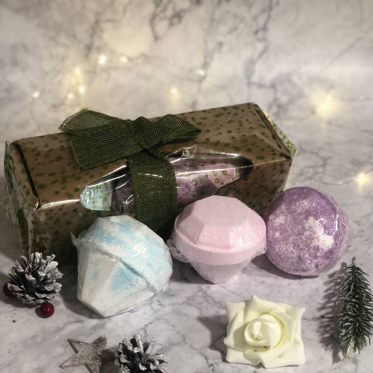 Gift set with three colorful bath bombs, wrapped in a brown polka-dotted box tied with a green ribbon, surrounded by pine cones, berries, and a decorative white flower on marble surface.