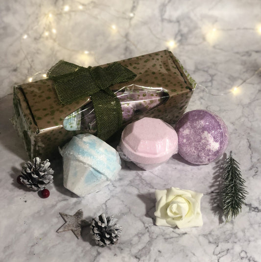 Bath bombs rest on a marbled surface, accompanied by a gift box tied with a green ribbon. Pinecones, a white rose, and a small pine branch decorate the scene, with soft fairy lights in the background.