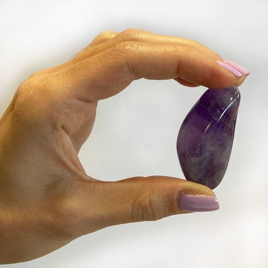 A hand holds an amethyst stone between the thumb and forefinger. The background is white.