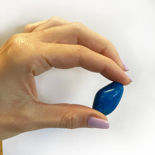 A hand holds a polished, dark blue stone between the thumb and forefinger. The hand has pink painted nails. The background is white.