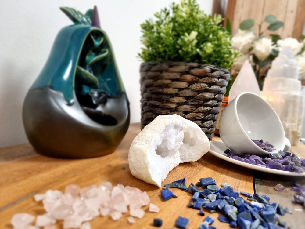 A ceramic, pear-shaped incense holder sits with descending smoke. Crystals and a tipped tea cup with amethyst pebbles surround a wooden surface with a potted plant.