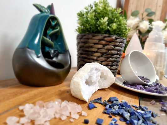A ceramic, pear-shaped incense holder sits with descending smoke. Crystals and a tipped tea cup with amethyst pebbles surround a wooden surface with a potted plant.